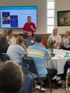 Hank Prince speaking at the Aphasia Awareness Dinner 2023. The screen shows two books that Hank recommends for reading. 